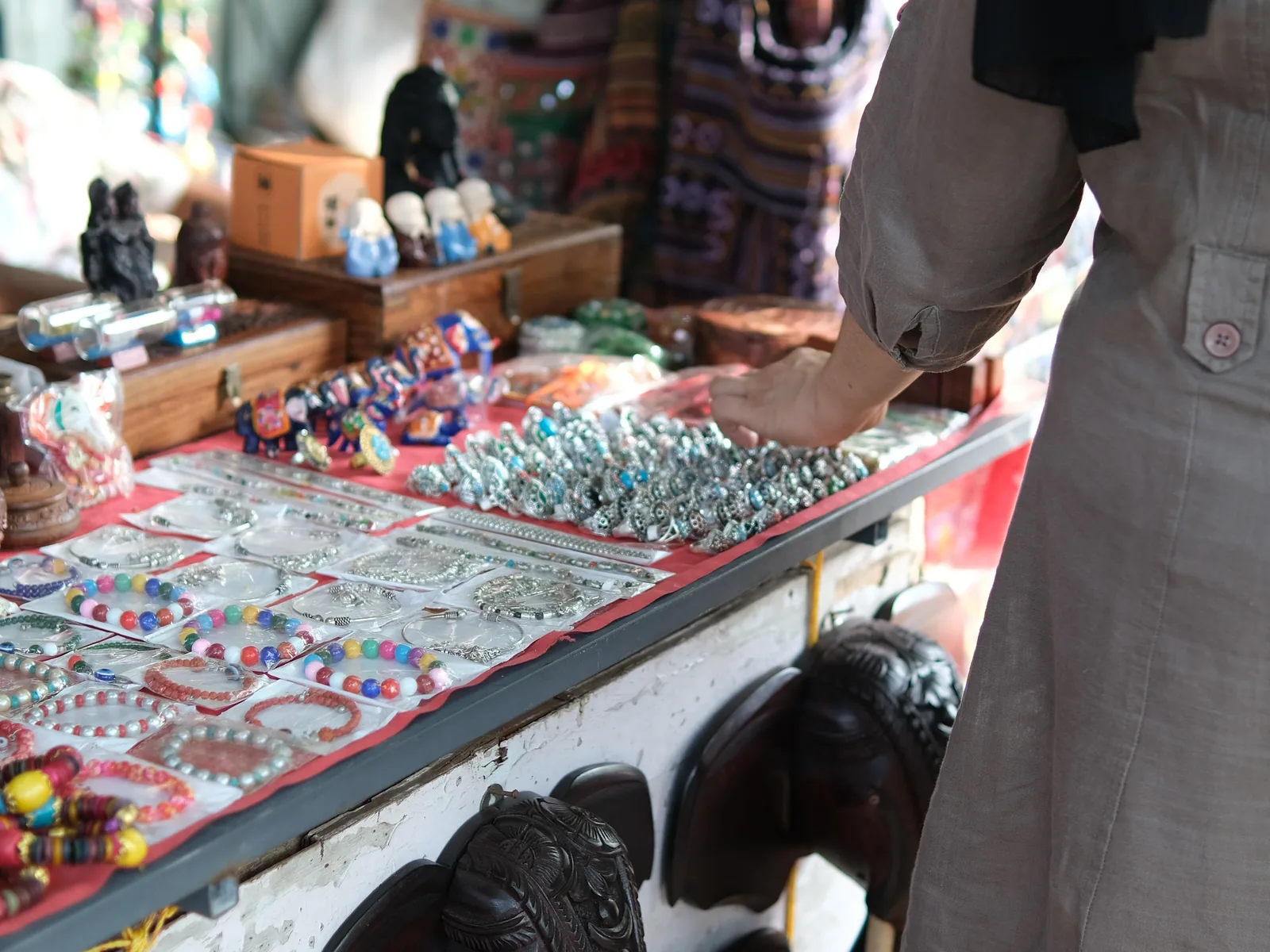 Close-up of a person's arm in a grey shirt reaching toward silver beads and jewelry at a market stall, with carved wooden elephants and colorful beaded necklaces displayed nearby.